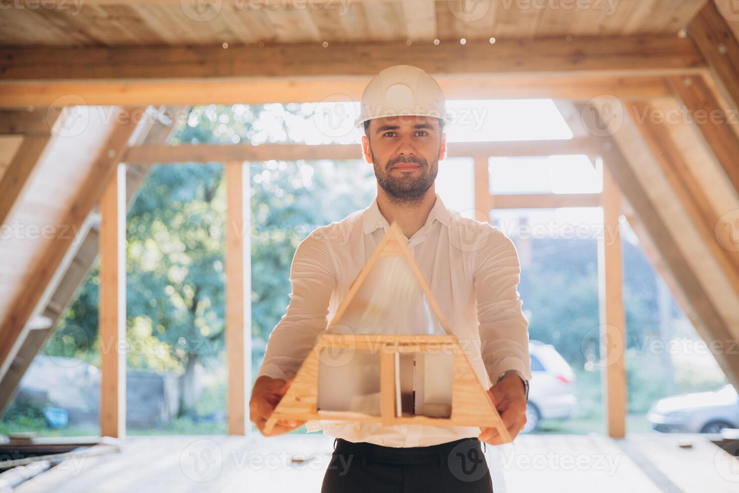 Man builder. Contractor for construction company. Architect in unfinished building. Builder in wooden modular house under construction. Male engineer holding a model of a miniature wooden house photo