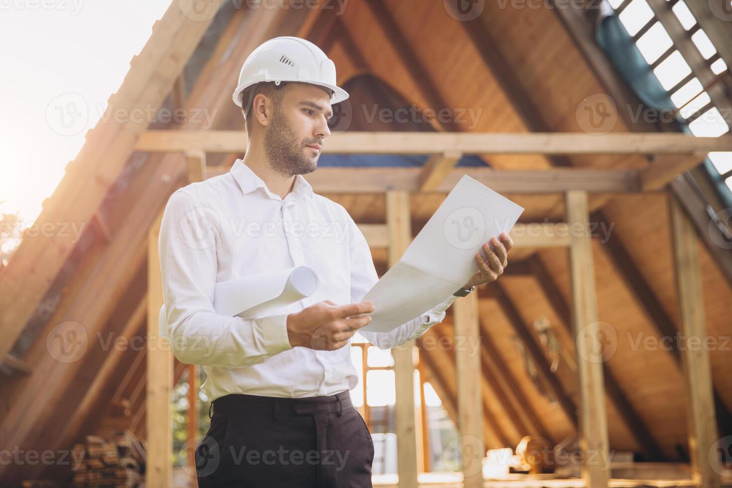 Architect wooden buildings. Man stands under frame wooden house. Man architect studies documentation or blueprints. Guy in protective helmet at construction site. Architect under timber framed house photo