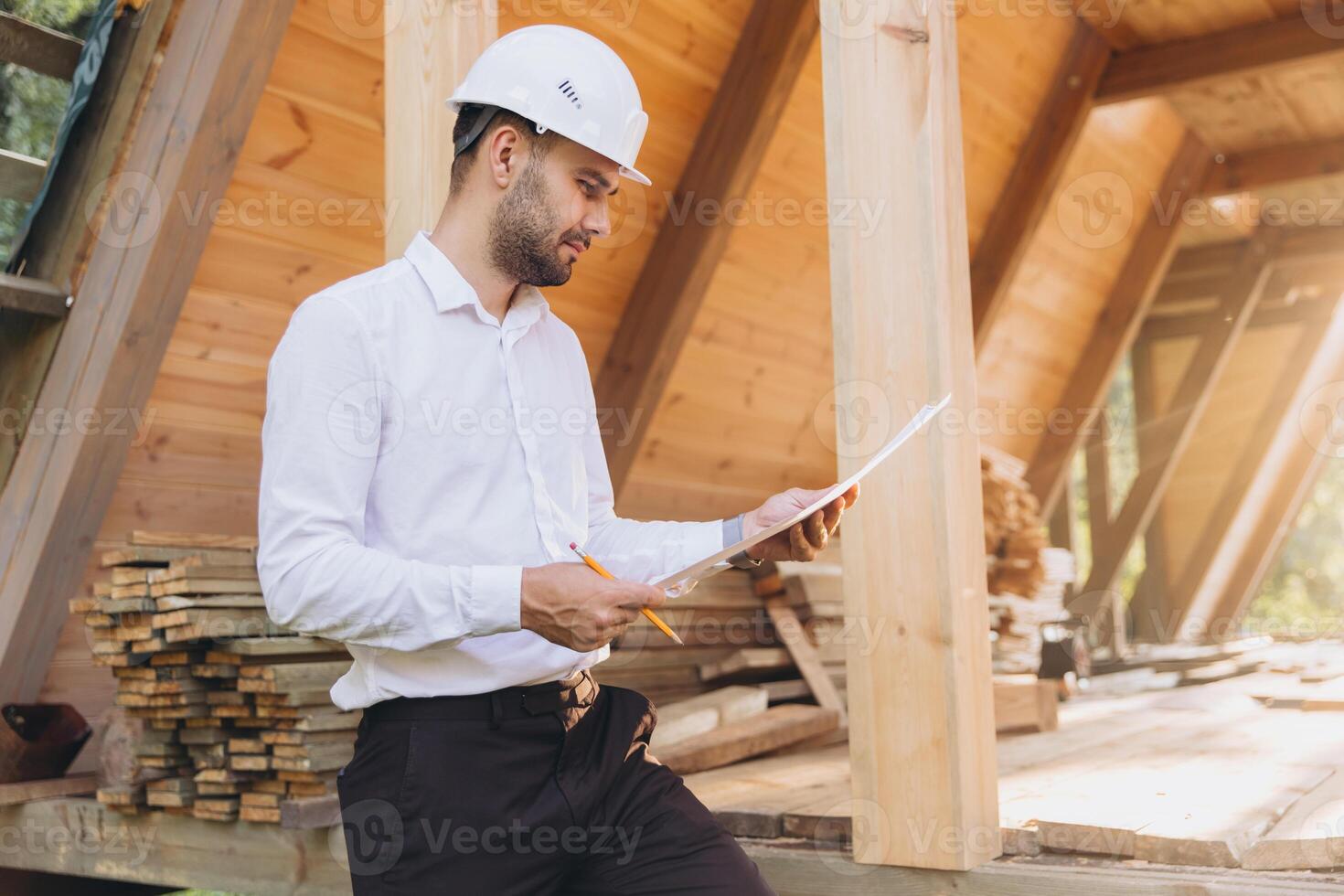 Construction engineer in white helmet. Developer with construction documentation on the background of wooden building under construction. Portrait of smiling architect near eco friendly house. photo