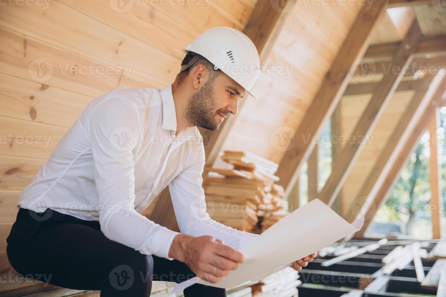 Architect wooden buildings. Man stands under frame wooden house. Man architect studies documentation or blueprints. Guy in protective helmet at construction site. Architect under timber framed house photo