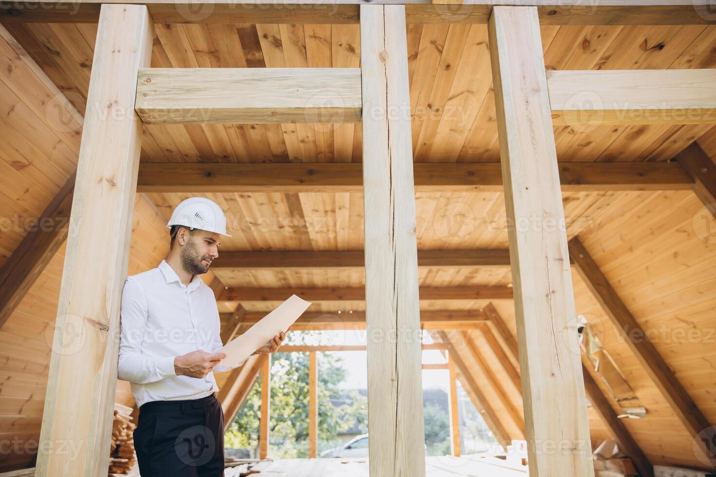 Architect wooden buildings. Man stands under frame wooden house. Man architect studies documentation or blueprints. Guy in protective helmet at construction site. Architect under timber framed house photo