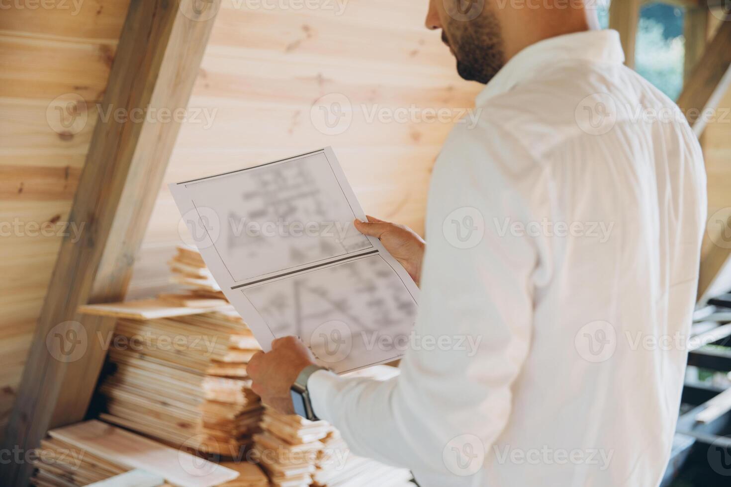 Architect wooden buildings. Man stands under frame wooden house. Man architect studies documentation or blueprints. Guy in protective helmet at construction site. Architect under timber framed house photo