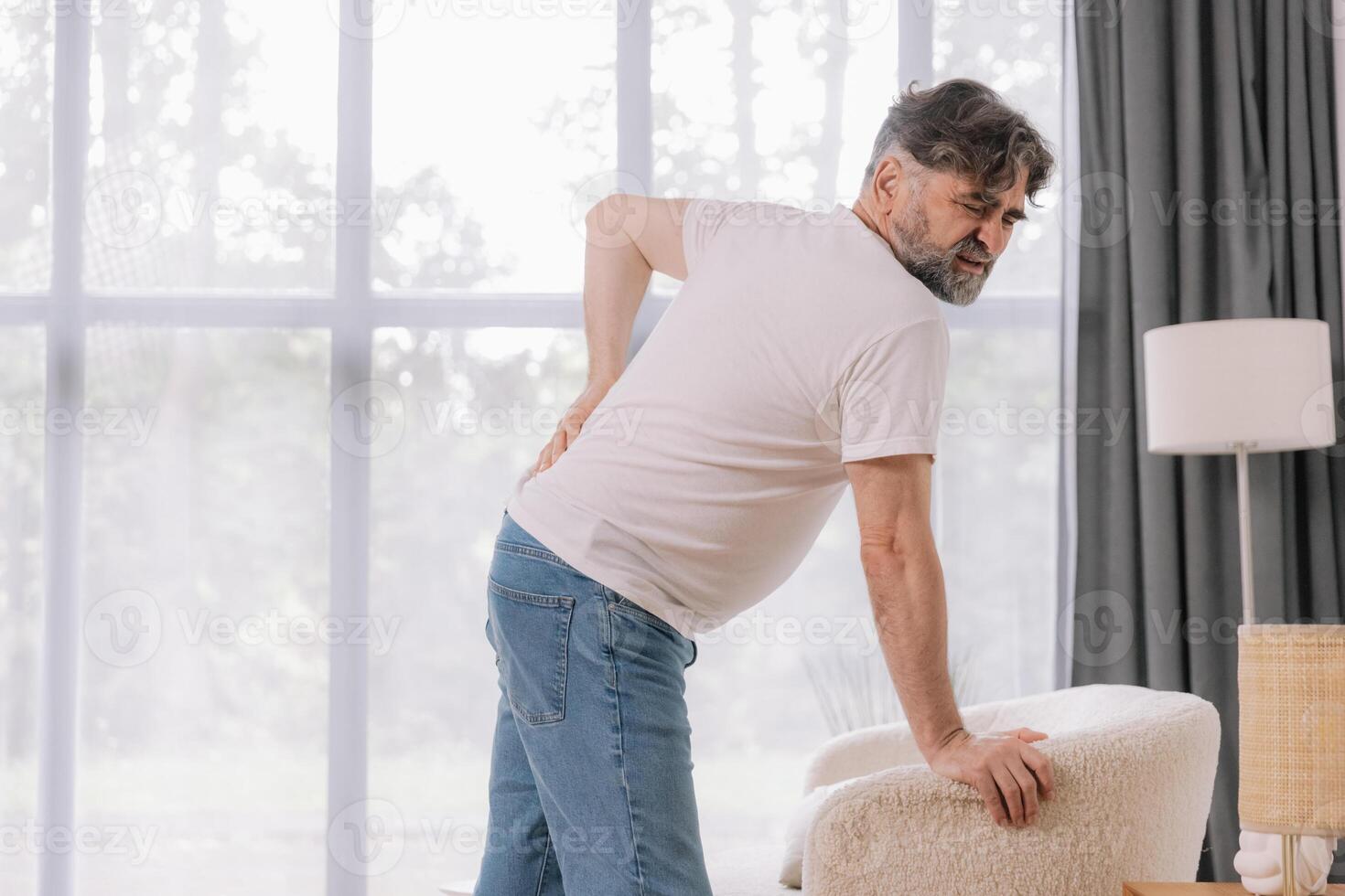 Rear view of senior man suffering from back pain on sofa at home, copy space for your advertising photo