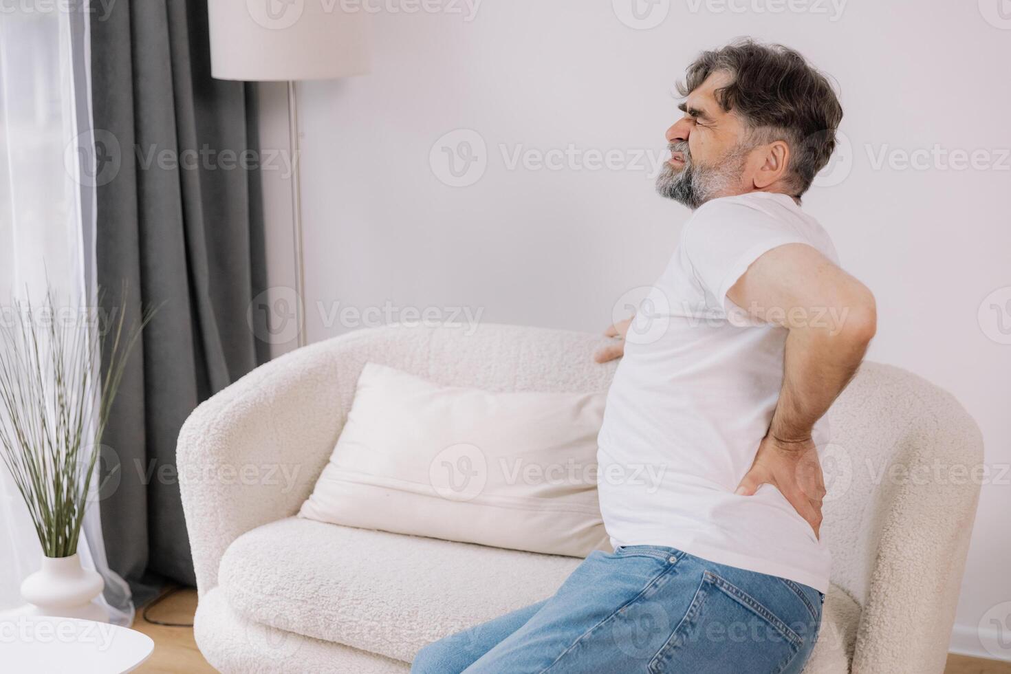Rear view of senior man suffering from back pain on sofa at home, copy space for your advertising photo