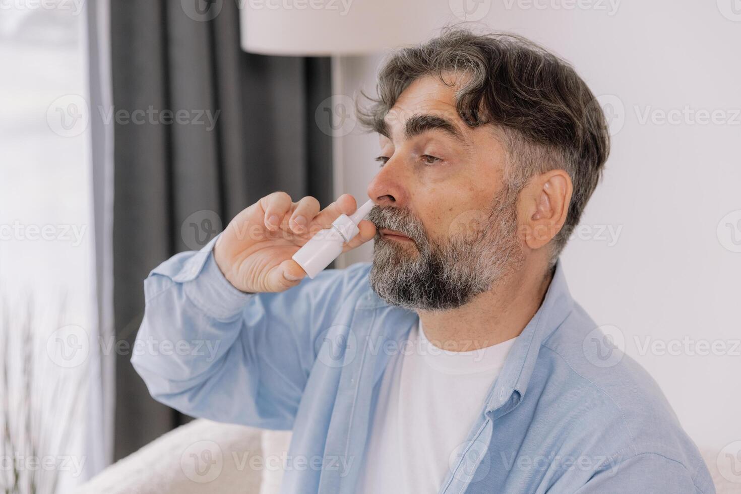 Elderly mature sick man in blanket suffering flu and cold using nasal spray at home. Rhinitis, medicine and healthcare concept photo