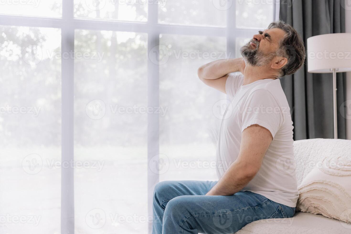 A senior man is sitting indoors, visibly suffering from neck pain. He is holding his neck with a pained expression, indicating discomfort. photo