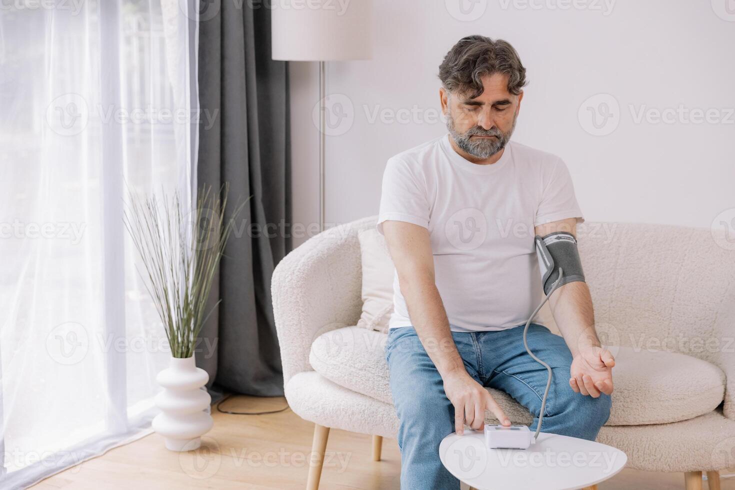 Handsome grey-haired senior man in casual outfit sitting on couch at home, checking blood pressure on his own. Pensioner using modern tonometer, experiencing hypertension, copy space photo