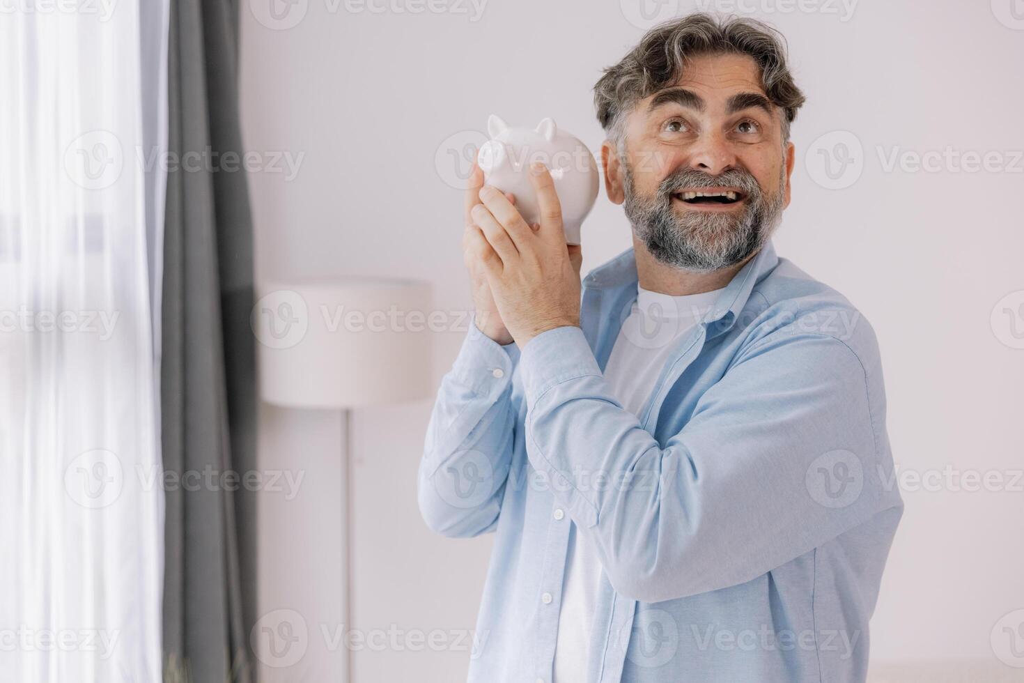 Senior man with a beard shows a white piggy bank suggesting themes of savings, retirement, and financial planning with copy space photo