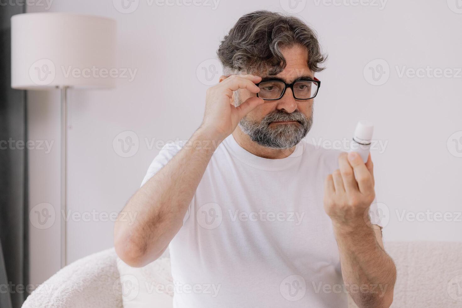 Senior man in glasses with poor vision reads description and instructions for medicine at home, self-care concept, elderly people, copy space, mock up photo