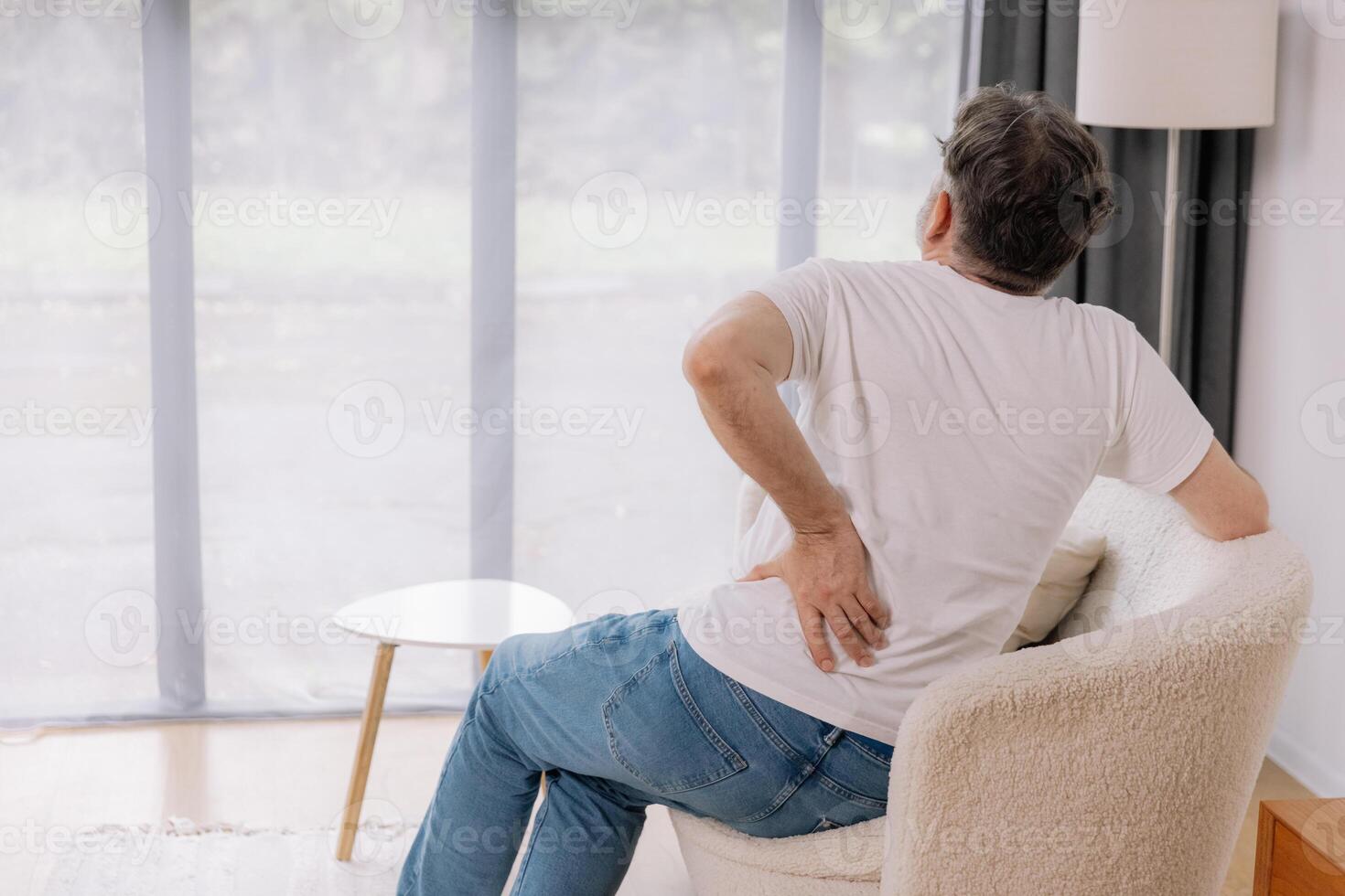 Rear view of senior man suffering from back pain on sofa at home, copy space for your advertising photo