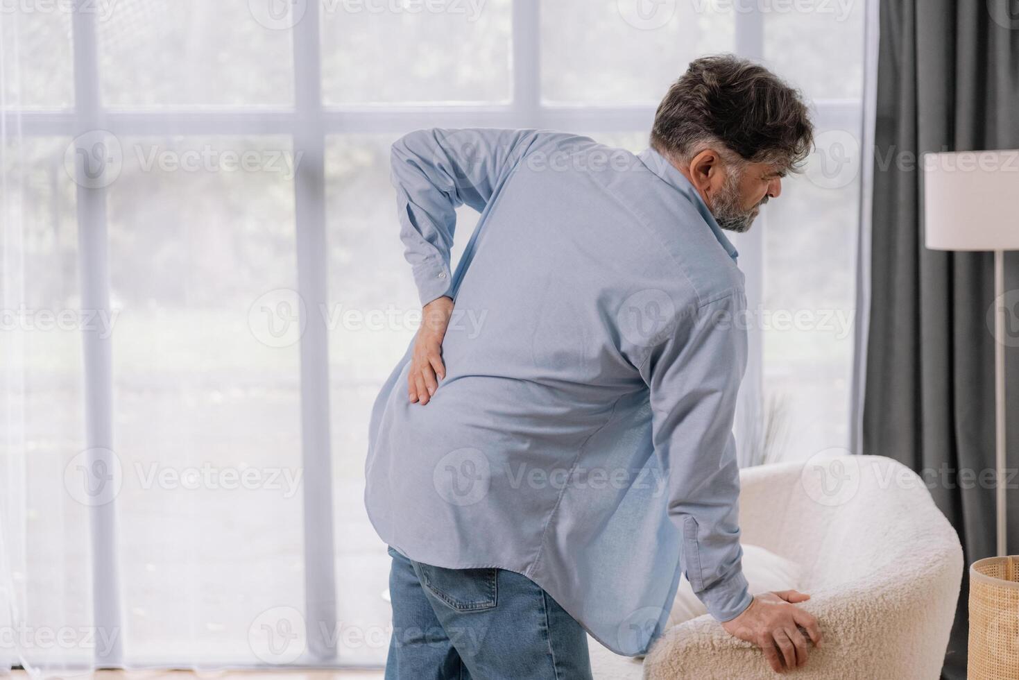 Rear view of senior man suffering from back pain on sofa at home, copy space for your advertising photo