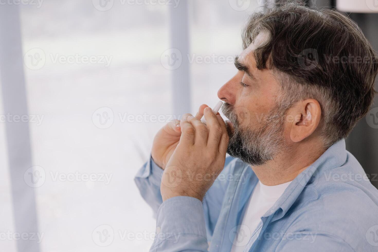 Elderly mature sick man in blanket suffering flu and cold using nasal spray at home. Rhinitis, medicine and healthcare concept photo