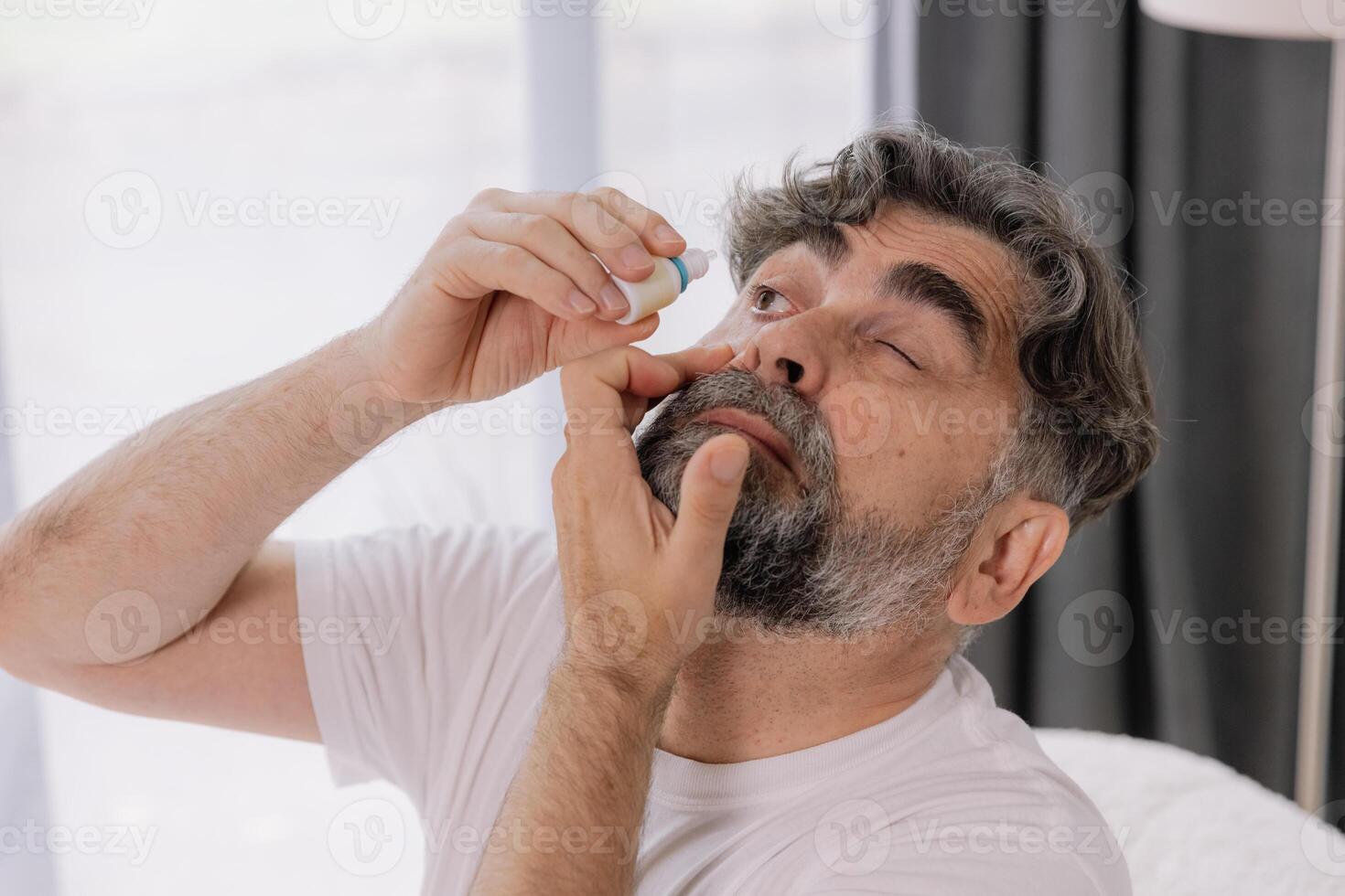 Close-up photo of an older gray-faced man who drips eye drops with medicine for pain and fatigue, sitting on the couch at home.