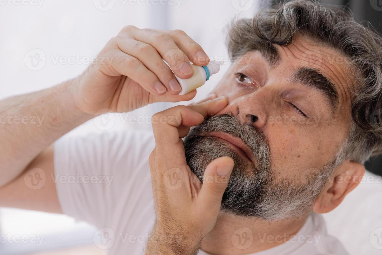 Close-up photo of an older gray-faced man who drips eye drops with medicine for pain and fatigue, sitting on the couch at home.
