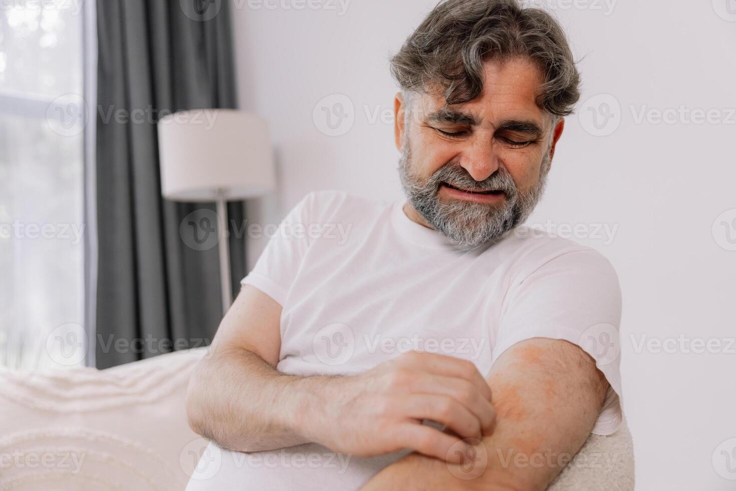 Senior man scratching forearm sitting on living room at home. Allergy symptom. Copy space photo