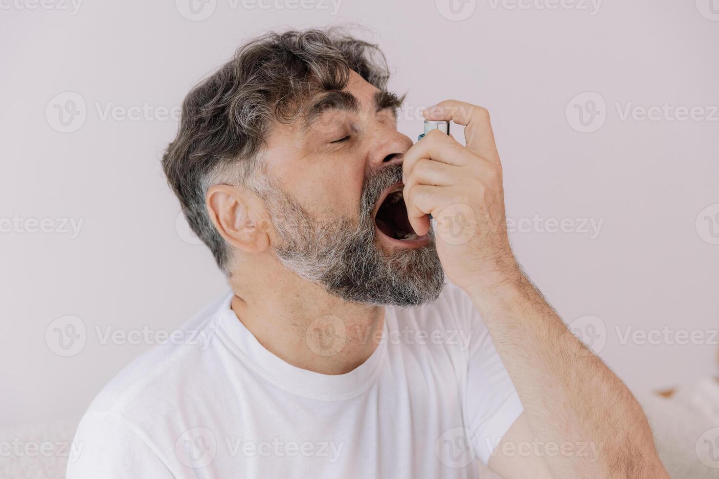 Sick grey-haired senior man touching his chest and using asthma inhaler for allergies, grandfather suffering from asthma attack at home, difficulties with breathing, closeup shot, copy space photo