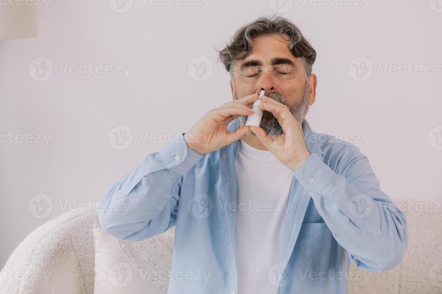 Elderly mature sick man in blanket suffering flu and cold using nasal spray at home. Rhinitis, medicine and healthcare concept photo