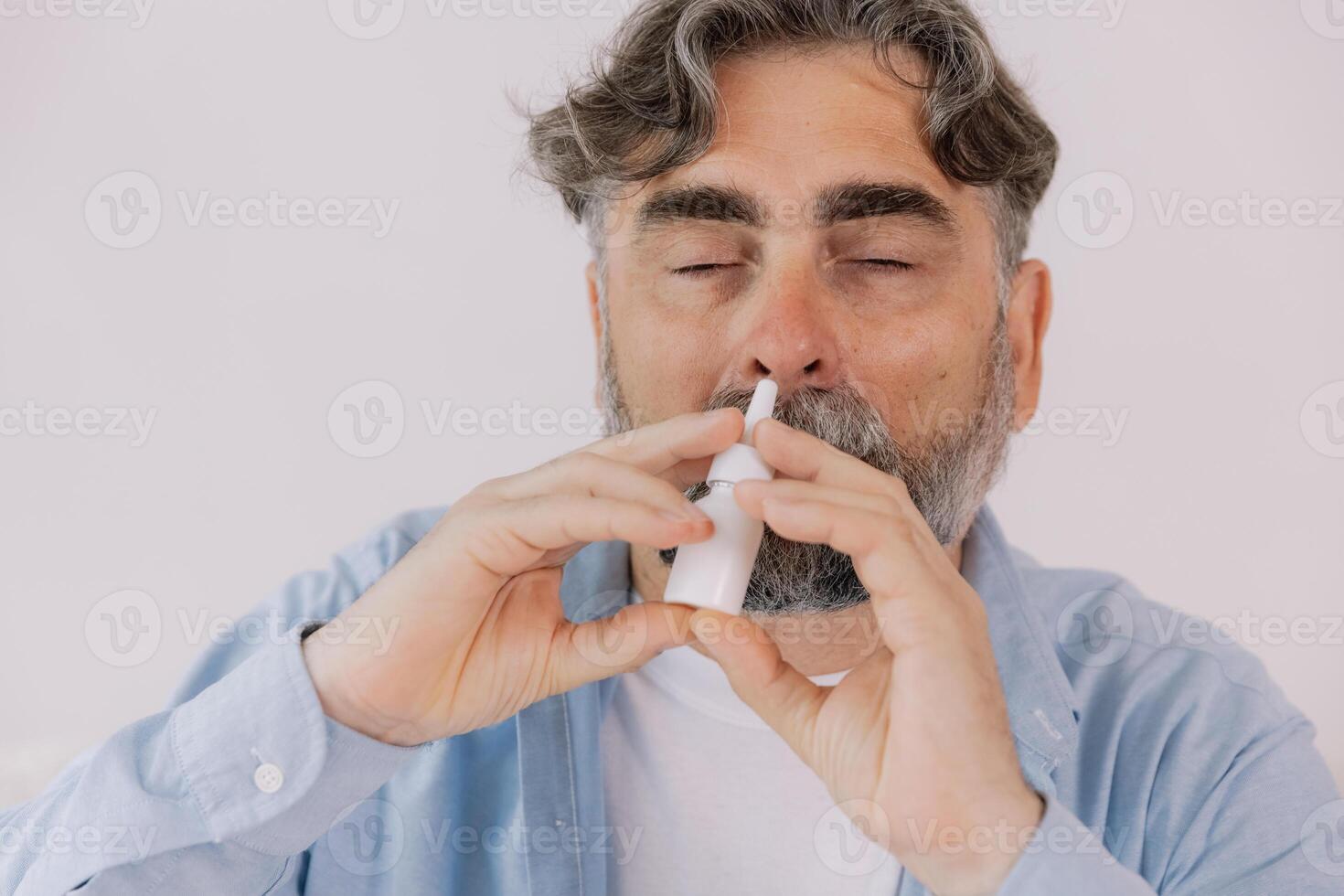 Elderly mature sick man in blanket suffering flu and cold using nasal spray at home. Rhinitis, medicine and healthcare concept photo