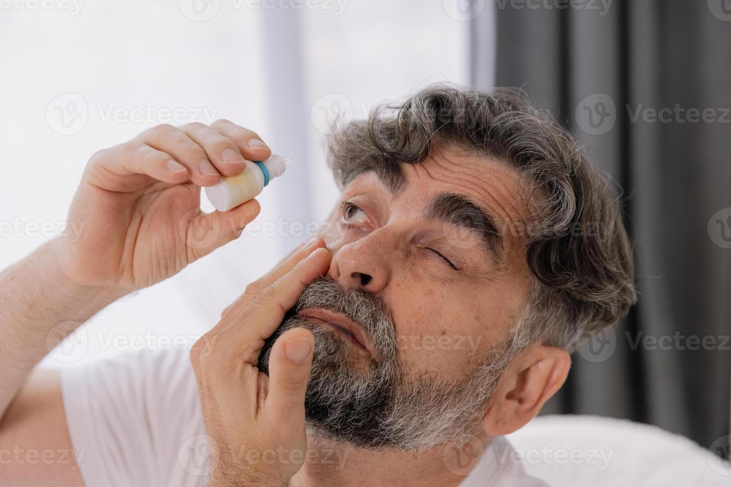 Close-up photo of an older gray-faced man who drips eye drops with medicine for pain and fatigue, sitting on the couch at home.