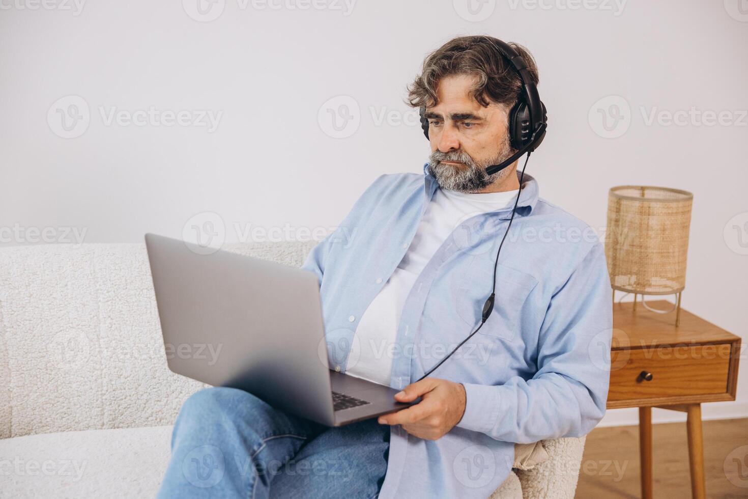 Positive grandfather using laptop computer and headset, sitting on couch. Senior man working from home, have online meeting, chat, copy space photo