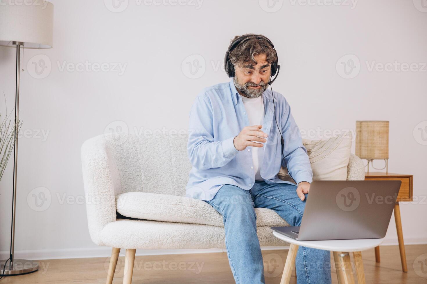 Positive grandfather using laptop computer and headset, sitting on couch. Senior man working from home, have online meeting, chat, copy space photo