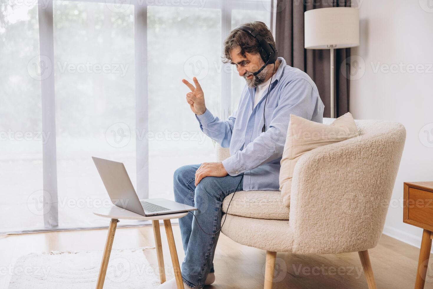 Positive grandfather using laptop computer and headset, sitting on couch. Senior man working from home, have online meeting, chat, copy space photo