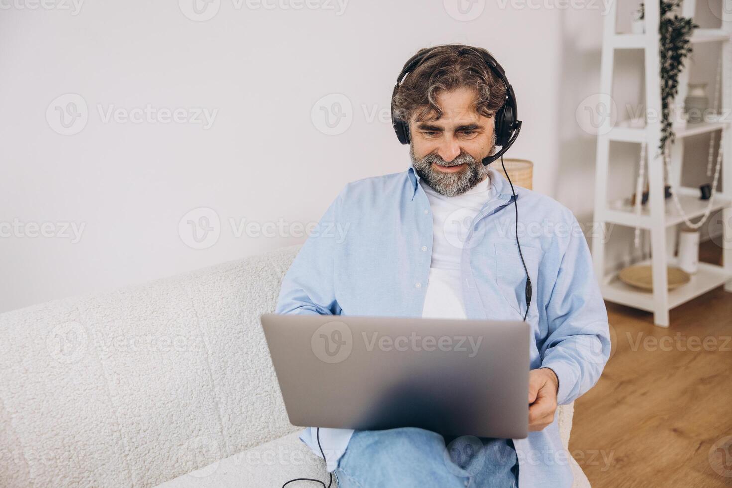 Positive grandfather using laptop computer and headset, sitting on couch. Senior man working from home, have online meeting, chat, copy space photo