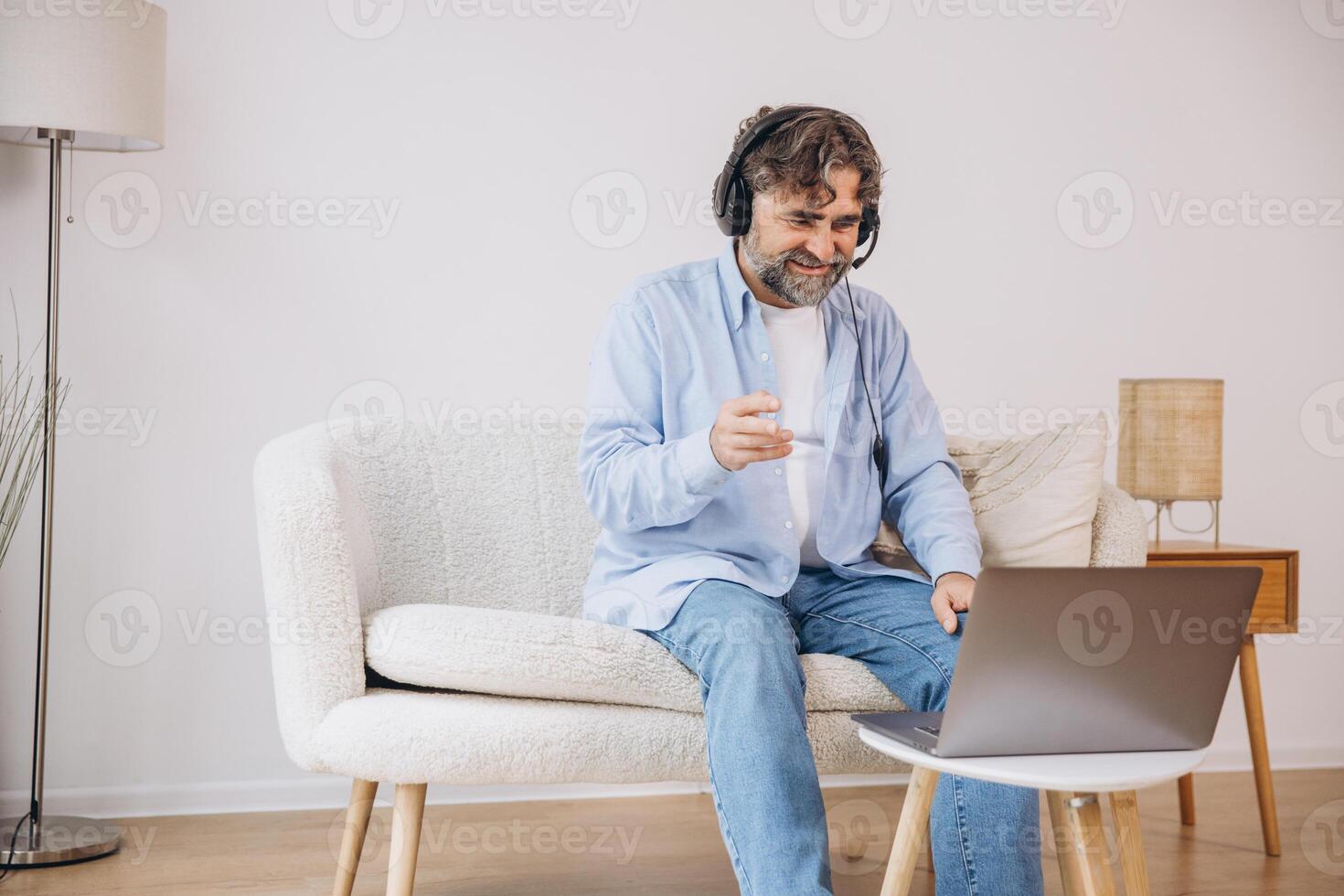 Positive grandfather using laptop computer and headset, sitting on couch. Senior man working from home, have online meeting, chat, copy space photo