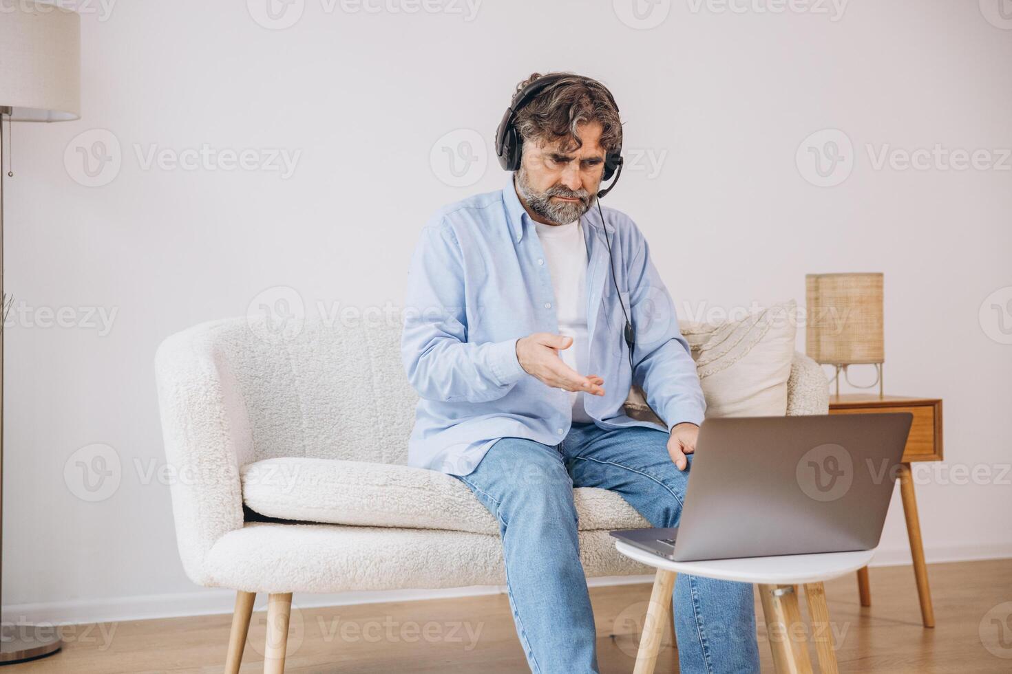 Positive grandfather using laptop computer and headset, sitting on couch. Senior man working from home, have online meeting, chat, copy space photo