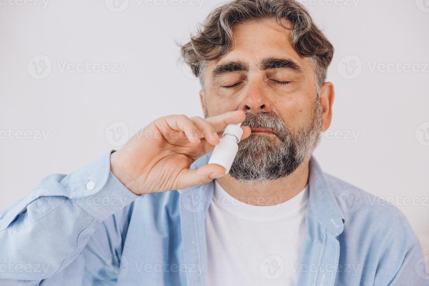 Elderly mature sick man in blanket suffering flu and cold using nasal spray at home. Rhinitis, medicine and healthcare concept photo