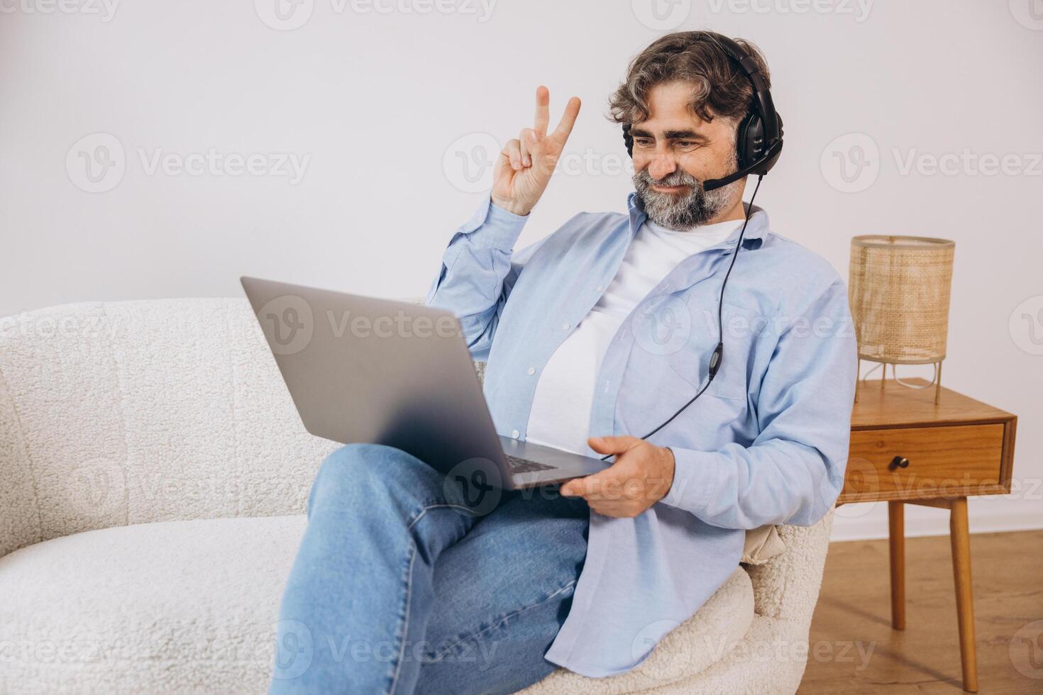 Positive grandfather using laptop computer and headset, sitting on couch. Senior man working from home, have online meeting, chat, copy space photo