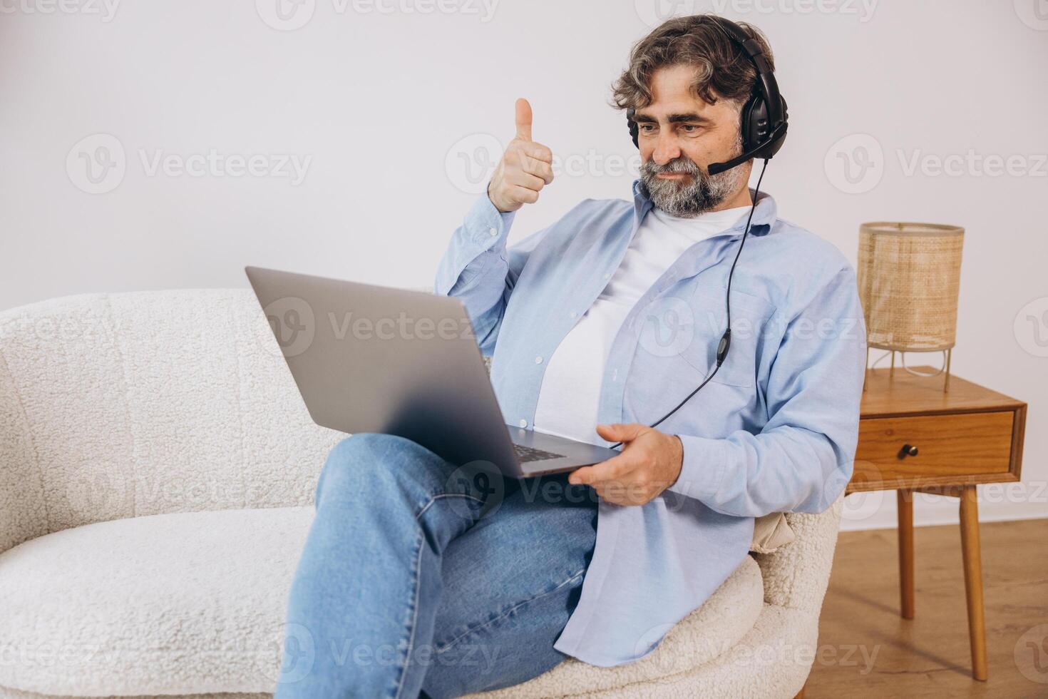 Positive grandfather using laptop computer and headset, sitting on couch. Senior man working from home, have online meeting, chat, copy space photo