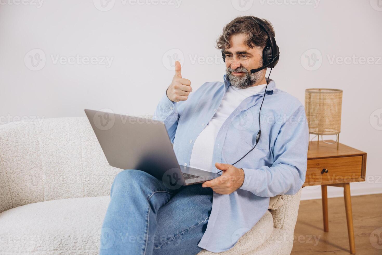 Positive grandfather using laptop computer and headset, sitting on couch. Senior man working from home, have online meeting, chat, copy space photo
