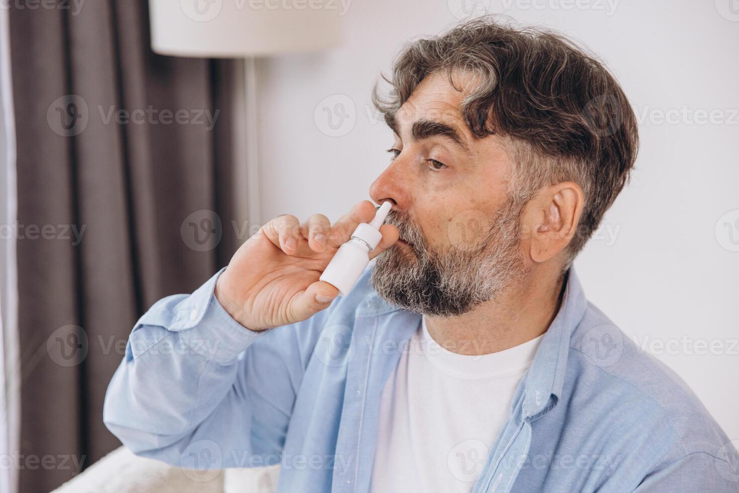 Elderly mature sick man in blanket suffering flu and cold using nasal spray at home. Rhinitis, medicine and healthcare concept photo