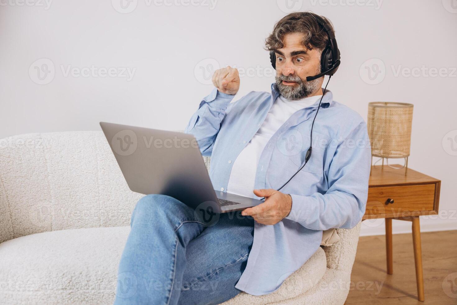 Positive grandfather using laptop computer and headset, sitting on couch. Senior man working from home, have online meeting, chat, copy space photo