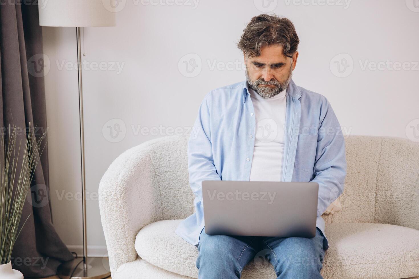 Smiling mature man using laptop sitting on armchair in living room. Happy confident senior male adult resting at home working on pc, typing on keyboard, copy space photo