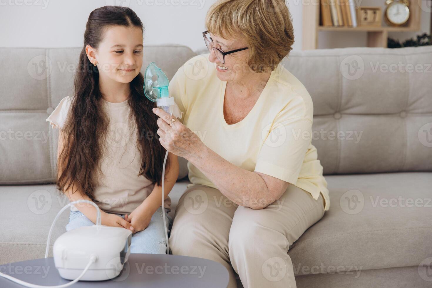 Little girl making inhalation with nebulizer with granny. Woman makes inhalation to a sick child while embracing her. Grandmother helping granddaughter with cold and flu to inahale nebuliser aerosol. photo