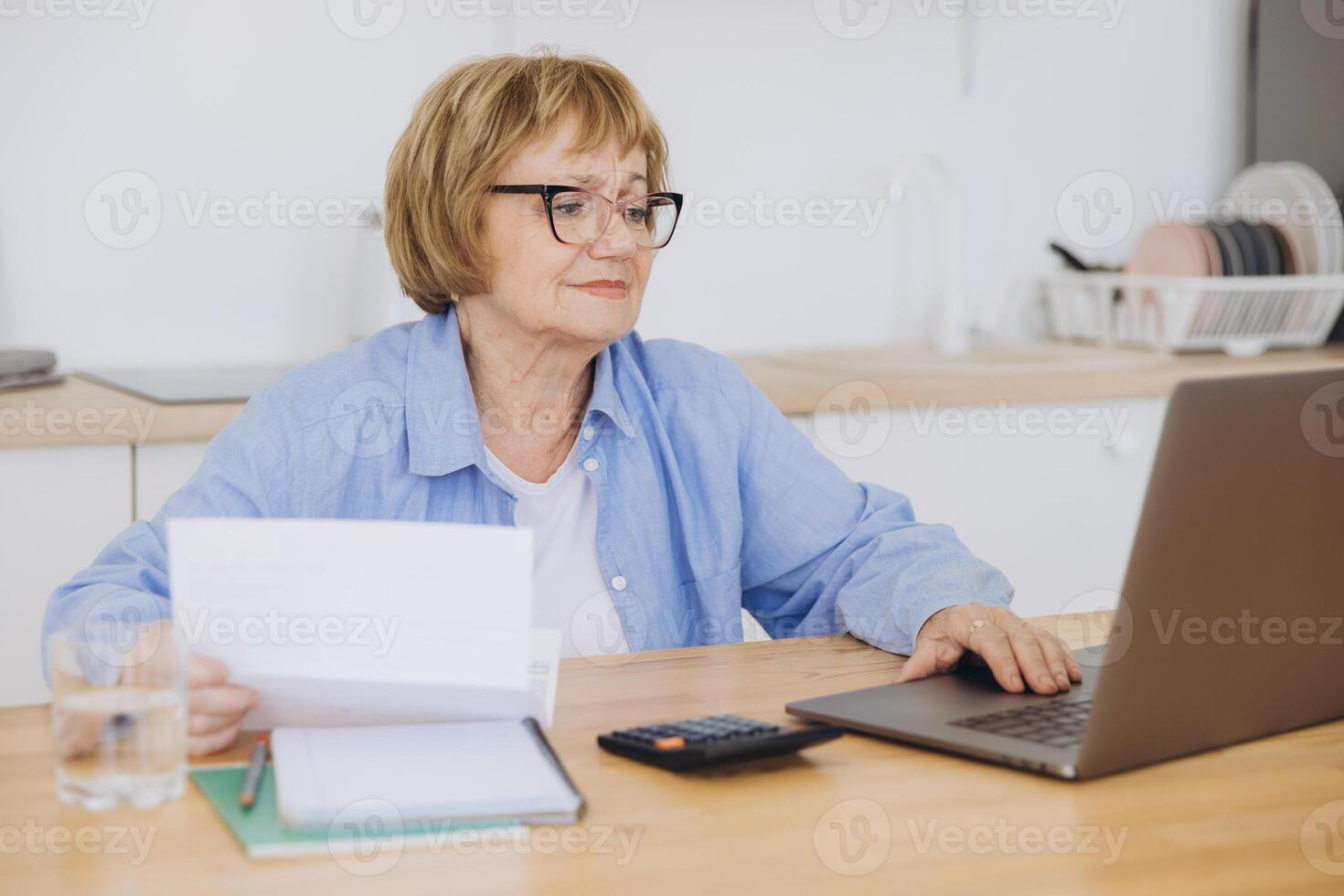 Senior business woman holding paper bill using calculator, old lady managing account finance, calculating money budget tax, planning banking loan debt pension payment sit at home kitchen table. photo