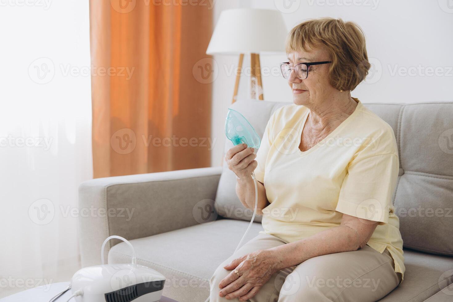 Sick senior woman has a viral infection or flu, labored breathing, asthma. An elderly female is using inhaler, nebulizer for self treatment sitting alone on the sofa at home photo