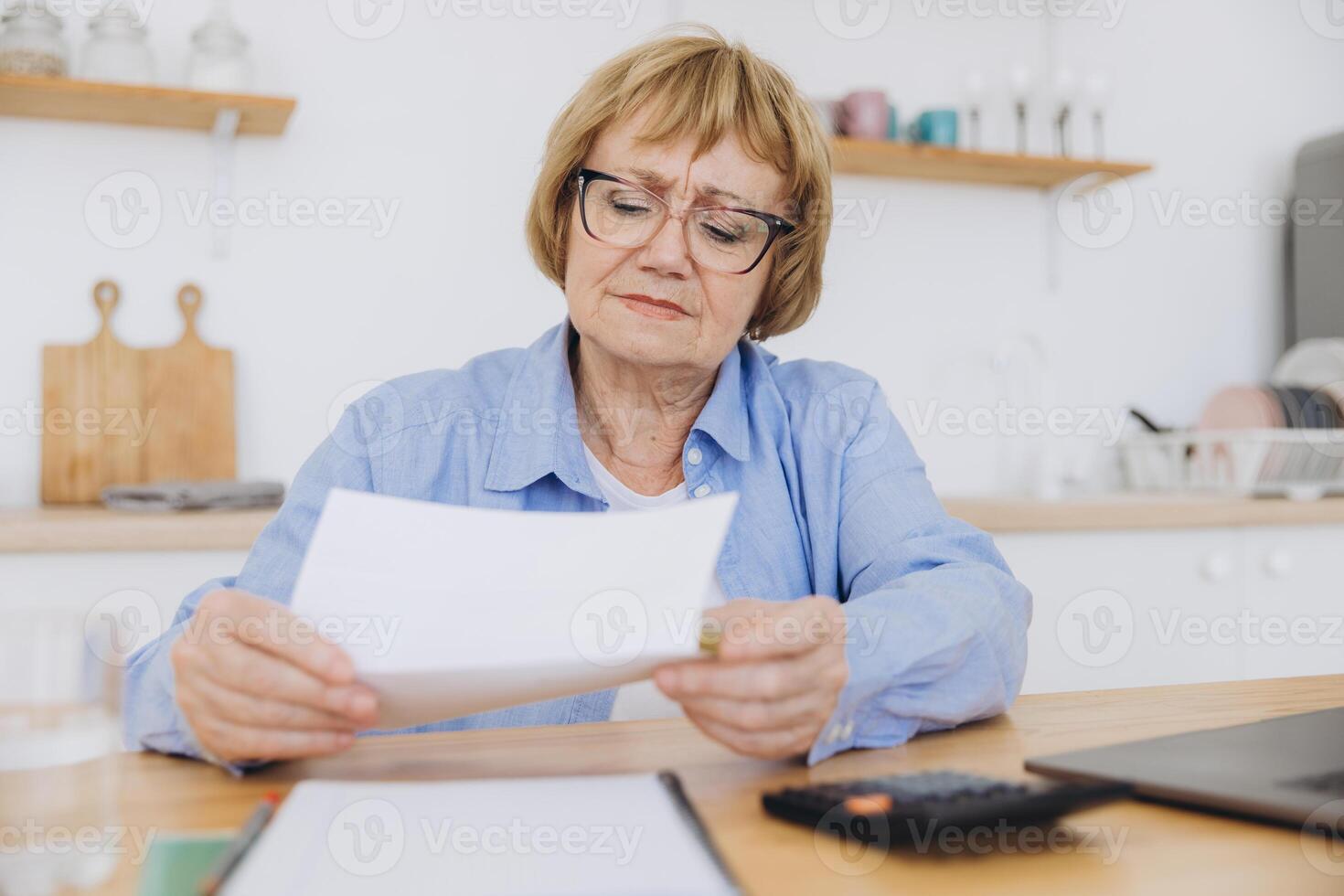 Shocked puzzled senior woman reading paper document, reviewing agreement terms, checking bills for payment at home table, getting angry about mistake, too high price, problem photo