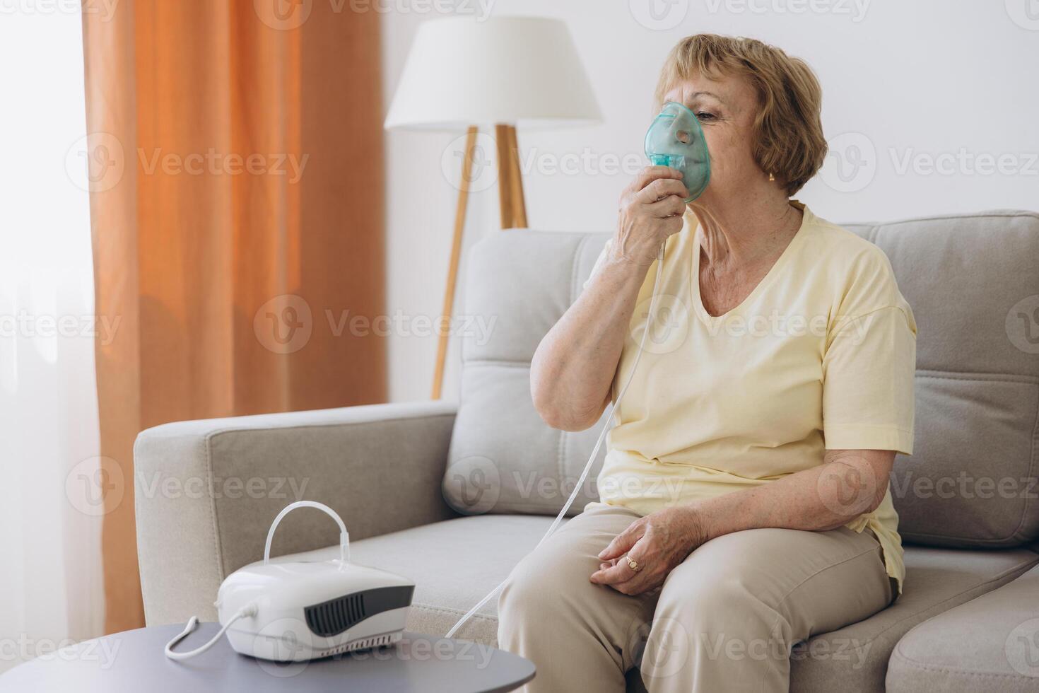 Sick senior woman has a viral infection or flu, labored breathing, asthma. An elderly female is using inhaler, nebulizer for self treatment sitting alone on the sofa at home photo