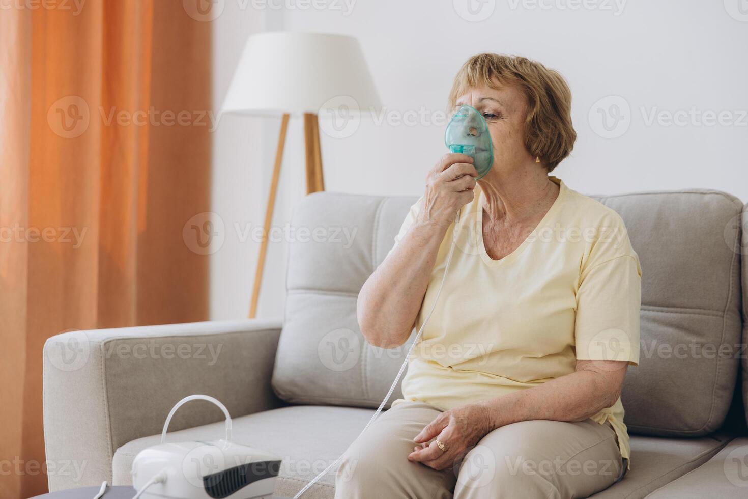 Sick senior woman has a viral infection or flu, labored breathing, asthma. An elderly female is using inhaler, nebulizer for self treatment sitting alone on the sofa at home photo