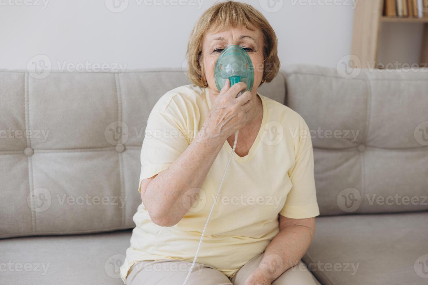 Sick senior woman has a viral infection or flu, labored breathing, asthma. An elderly female is using inhaler, nebulizer for self treatment sitting alone on the sofa at home photo