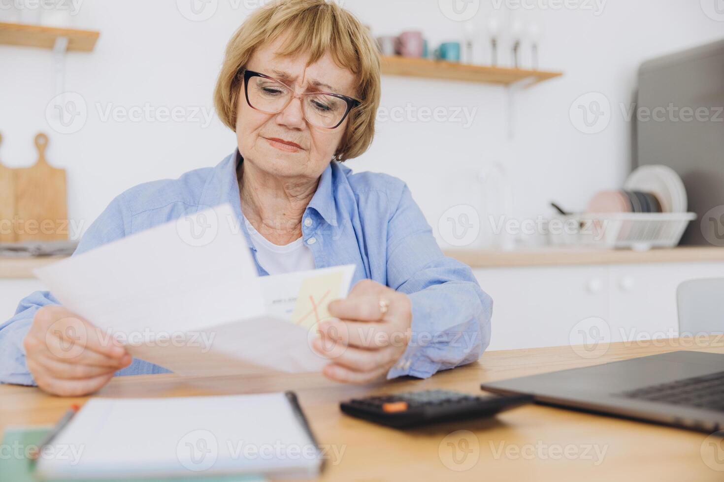 Shocked puzzled senior woman reading paper document, reviewing agreement terms, checking bills for payment at home table, getting angry about mistake, too high price, problem photo
