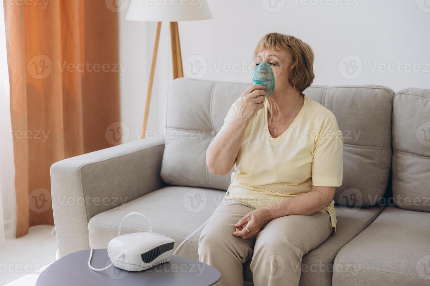 Sick senior woman has a viral infection or flu, labored breathing, asthma. An elderly female is using inhaler, nebulizer for self treatment sitting alone on the sofa at home photo