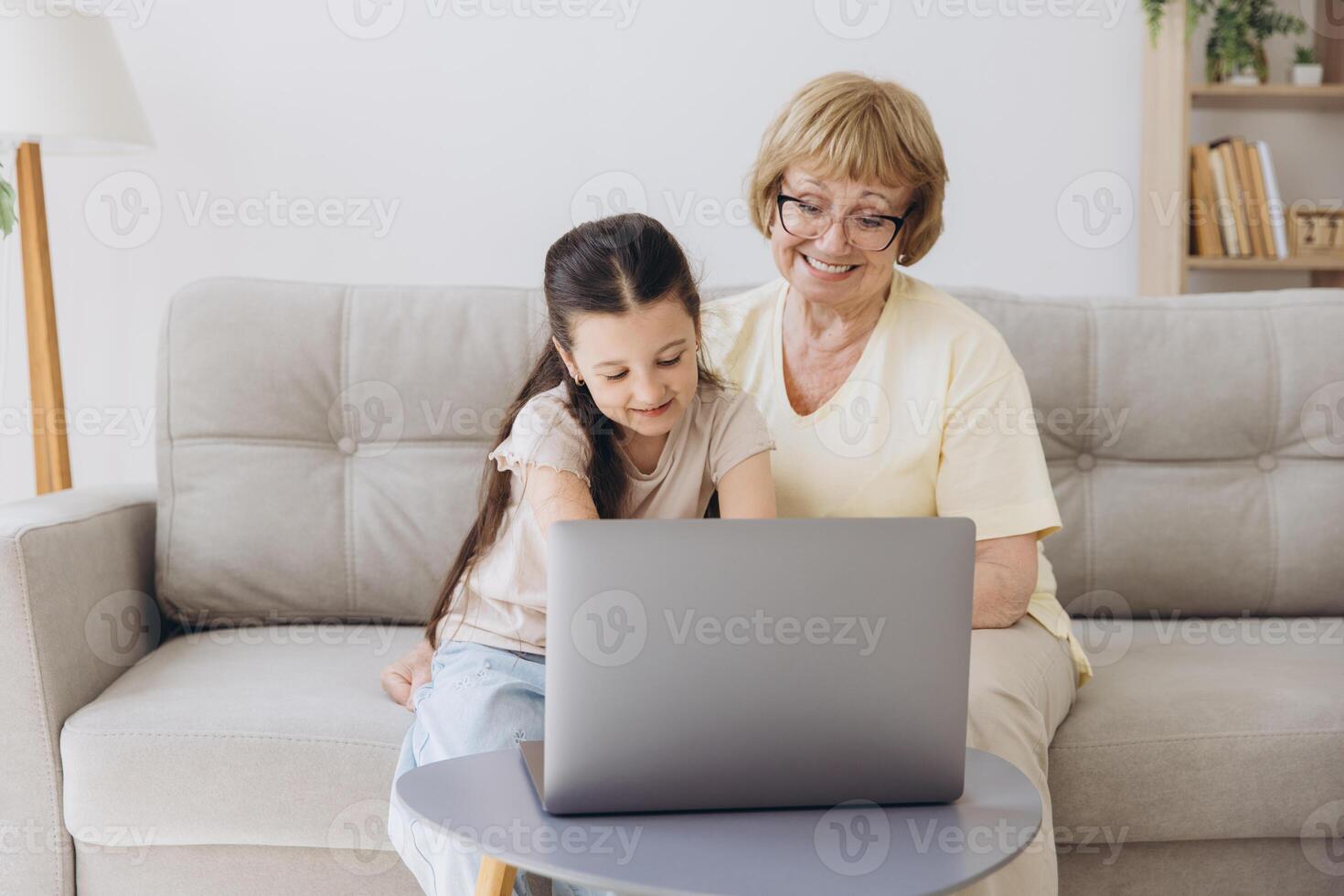 Multi generational family, grandma and granddaughter sit on couch with wireless computer, look at each other with tenderness and love, enjoy leisure on internet and time together, watch on-line movies photo