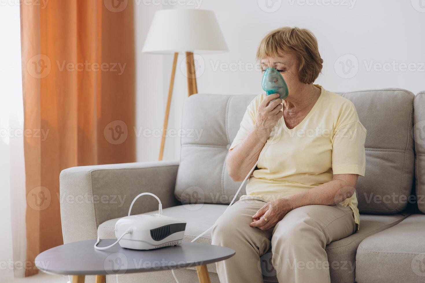 Sick senior woman has a viral infection or flu, labored breathing, asthma. An elderly female is using inhaler, nebulizer for self treatment sitting alone on the sofa at home photo