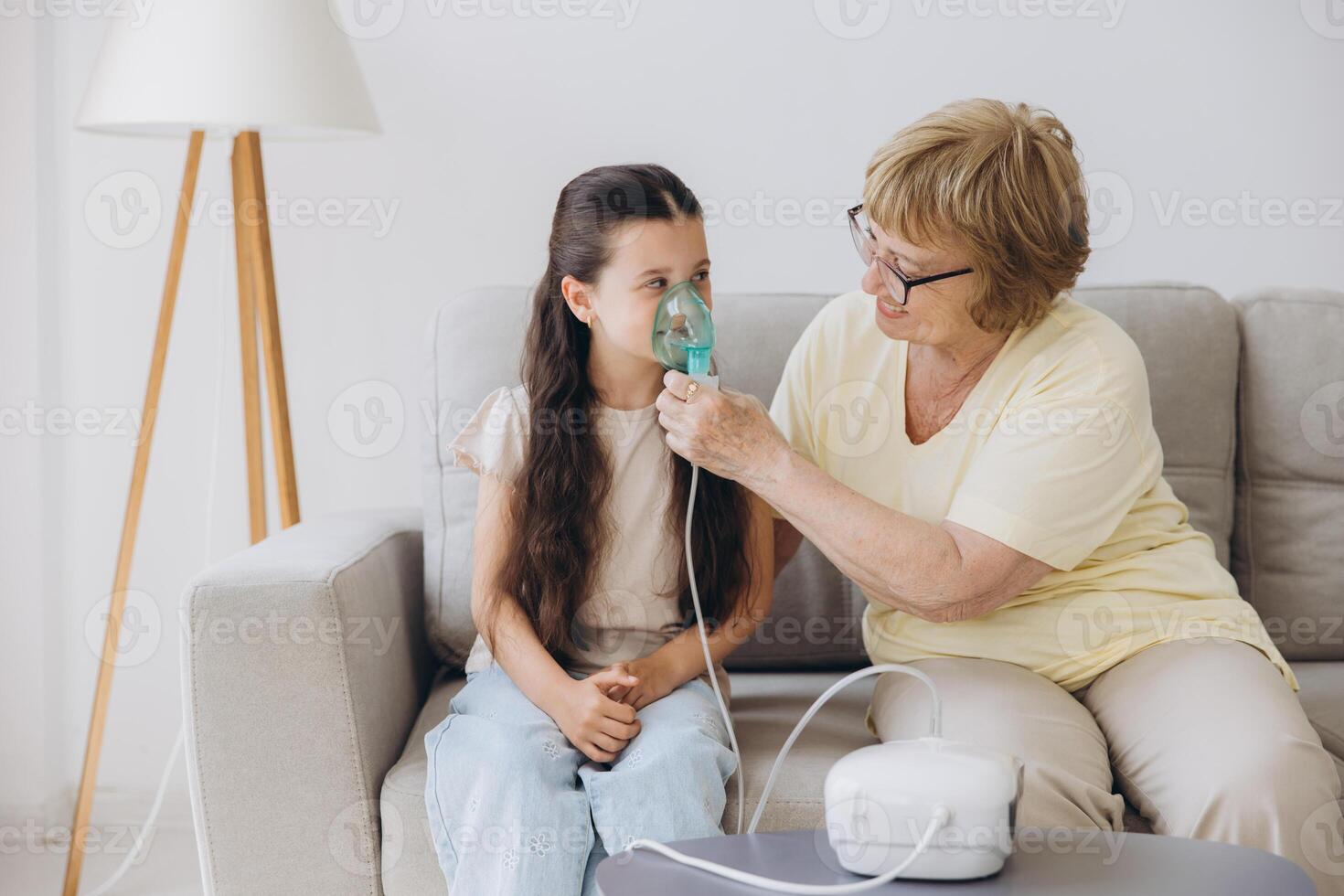 Little girl making inhalation with nebulizer with granny. Woman makes inhalation to a sick child while embracing her. Grandmother helping granddaughter with cold and flu to inahale nebuliser aerosol. photo