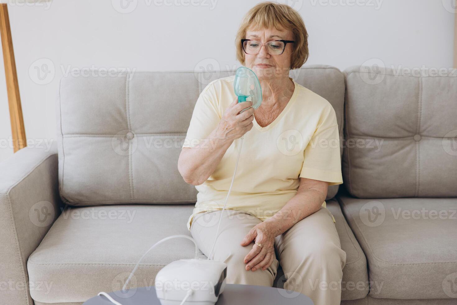 Sick senior woman has a viral infection or flu, labored breathing, asthma. An elderly female is using inhaler, nebulizer for self treatment sitting alone on the sofa at home photo