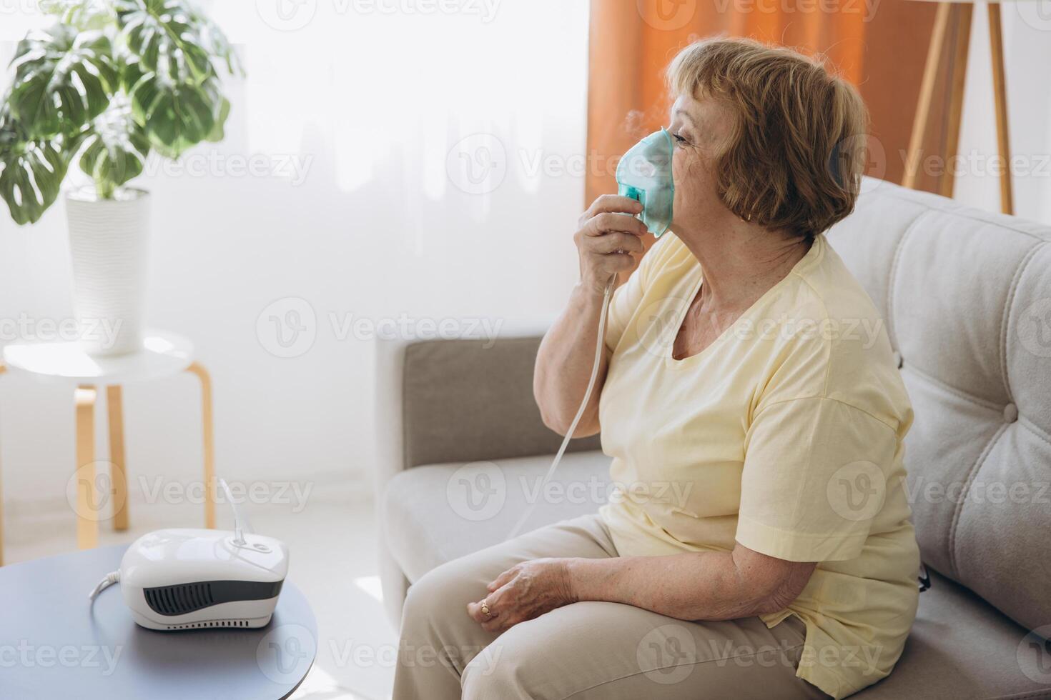 Sick senior woman has a viral infection or flu, labored breathing, asthma. An elderly female is using inhaler, nebulizer for self treatment sitting alone on the sofa at home photo